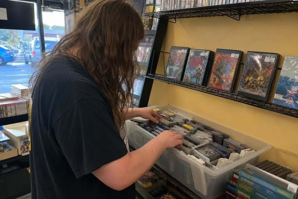 Person bläddrar bland lösa spelkassetter i en butik med retrospel, Dreamcast och Saturn syns i hyllan.