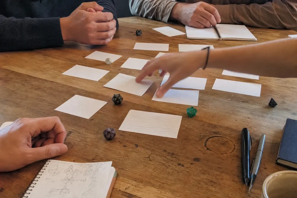 Flera händer samlade kring spelkort och tärningar på ett träbord under en session av Warrior Cats Clans of the Forest.