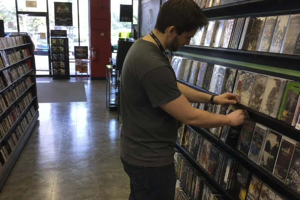 Butiksanställd fyller på hyllor med spel i en elektronikbutik inför GTA VI release, sent eftermiddagsljus genom fönstren.