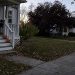 Halloween-utsmyckad veranda i skymning med utskurna pumpor och ett barn i utklädnad som går mot trottoaren.