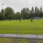 Svenska damfotbollsspelare tränar på gräsplan en mulen dag, fotograferat på avstånd från sidlinjen.